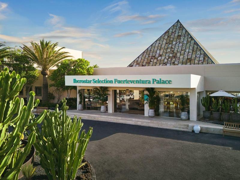 Ingresso di un resort con palme, cactus e un tetto a forma di piramide sotto un cielo azzurro.