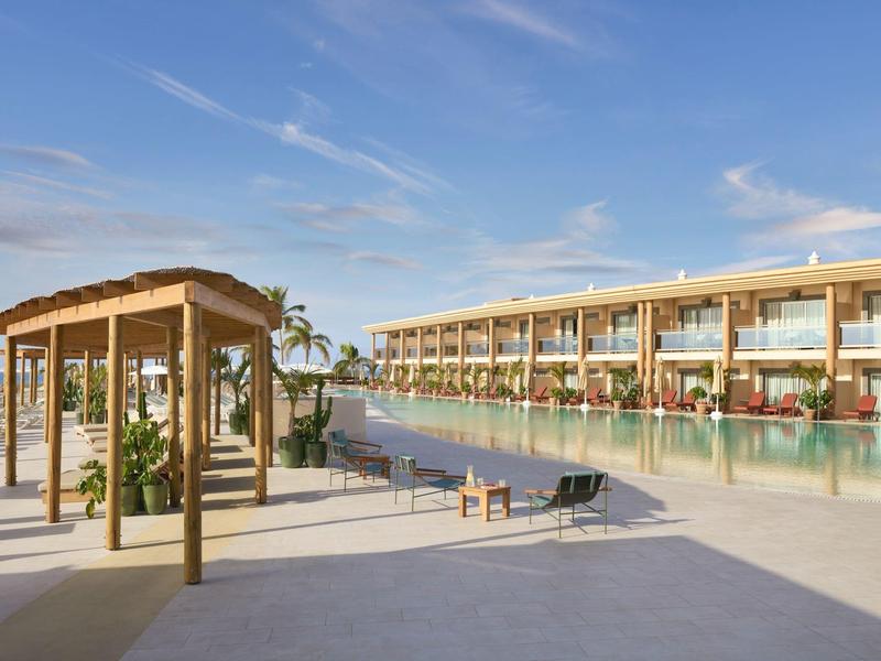 Complesso alberghiero moderno con piscina, ombrelloni e vista sul mare sotto un cielo limpido.