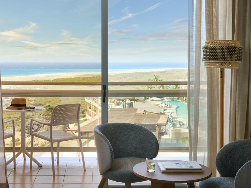 Camera d'hotel moderna con zona salotto e balcone con vista sulla piscina e sul mare.