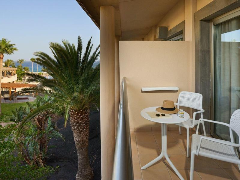 Balcone con tavolo bianco, due sedie e un cappello, vista su palme e area dell'hotel.