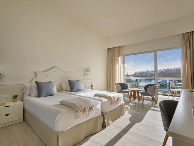 Camera d'hotel luminosa con due letti singoli, area salotto e vista sul mare attraverso grandi finestre.