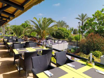 Terrasse de restaurant extérieure avec tables et chaises, entourée de végétation et palmiers sous un ciel bleu.