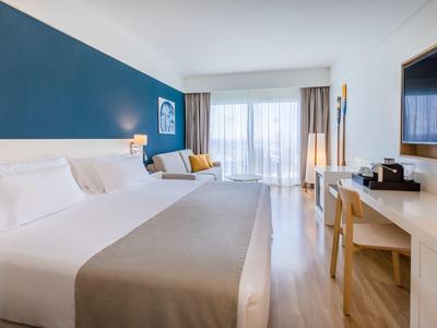 Chambre d'hôtel moderne avec grand lit, fauteuil, bureau, télévision et grande fenêtre lumineuse.