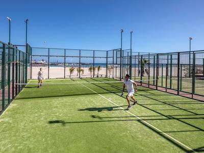 Zwei Personen spielen Padel-Tennis auf einem Outdoor-Feld mit grünem Kunstrasen unter blauem Himmel.