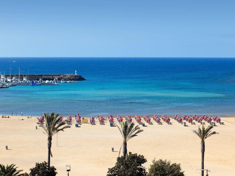 Plage tranquille avec parasols, mer bleue et palmiers en premier plan.