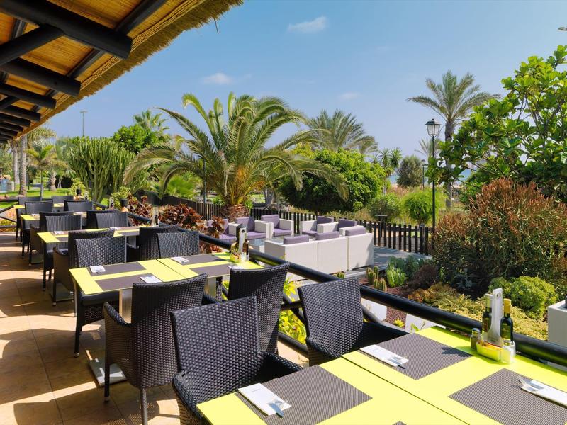 Terrasse de restaurant extérieure avec tables et chaises, entourée de végétation et palmiers sous un ciel bleu.