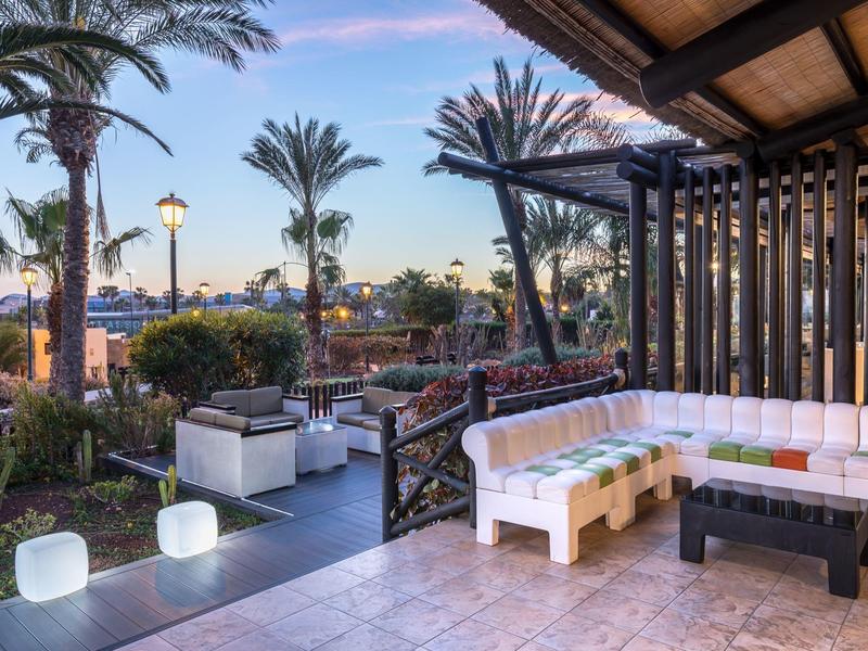 Terrasse d'hôtel avec mobilier extérieur face à un jardin avec palmiers au coucher du soleil.