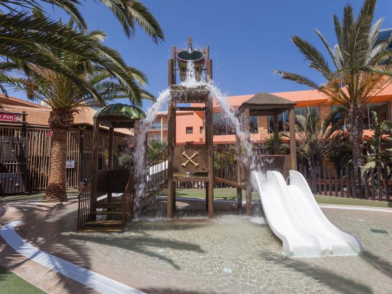 Aire de jeux avec toboggans blancs entourée de palmiers sous un ciel bleu.
