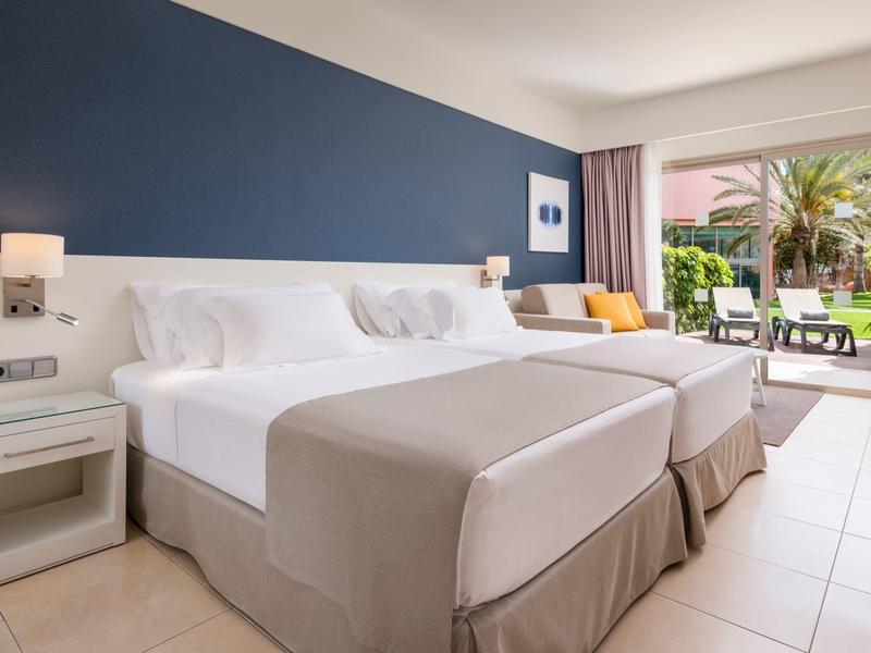 Chambre d'hôtel moderne avec deux lits, mur bleu, et terrasse avec vue sur jardin.