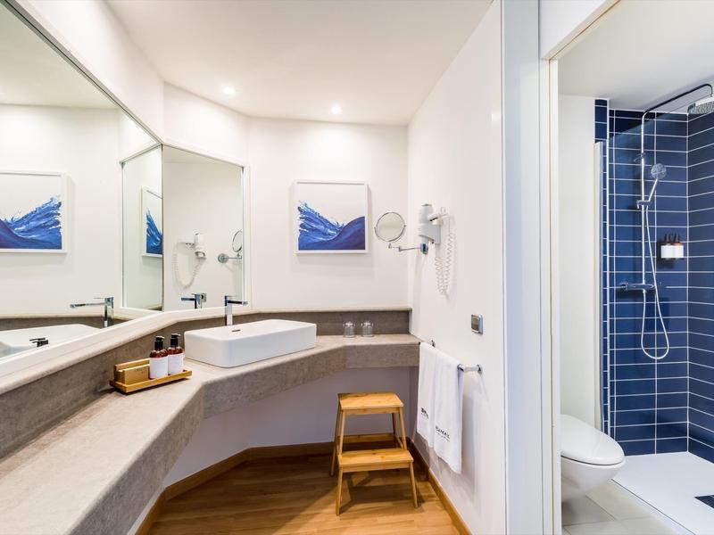 Salle de bain moderne avec lavabo, miroir, douche et tabouret en bois.