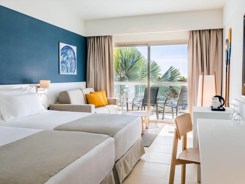 Chambre d'hôtel lumineuse avec deux lits, balcon, et vue sur la mer au loin.