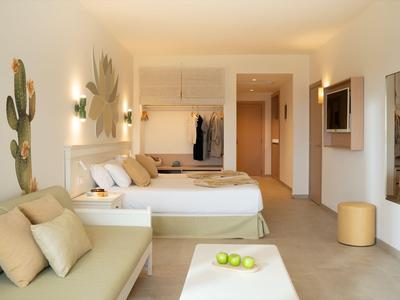 Nowoczesny pokój hotelowy z dwoma łóżkami, sofą i dekoracjami w beżowych, naturalnych tonach.