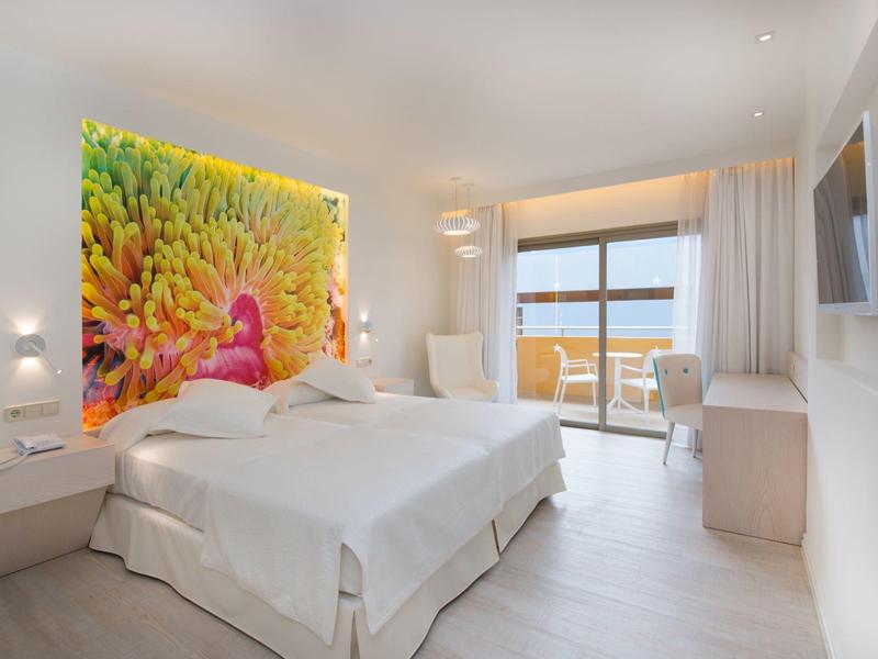 Nowoczesny pokój hotelowy z podwójnym łóżkiem, kolorową dekoracją ścienną i balkonem z widokiem na morze.