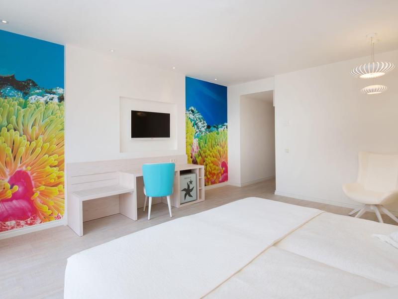 Jasny pokój hotelowy z kolorowymi obrazami na ścianach, biurkiem i krzesłem.