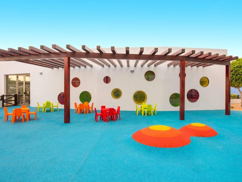 Kolorowy teren na zewnątrz z stołami, krzesłami i placem zabaw przed białą ścianą pod pergolą.