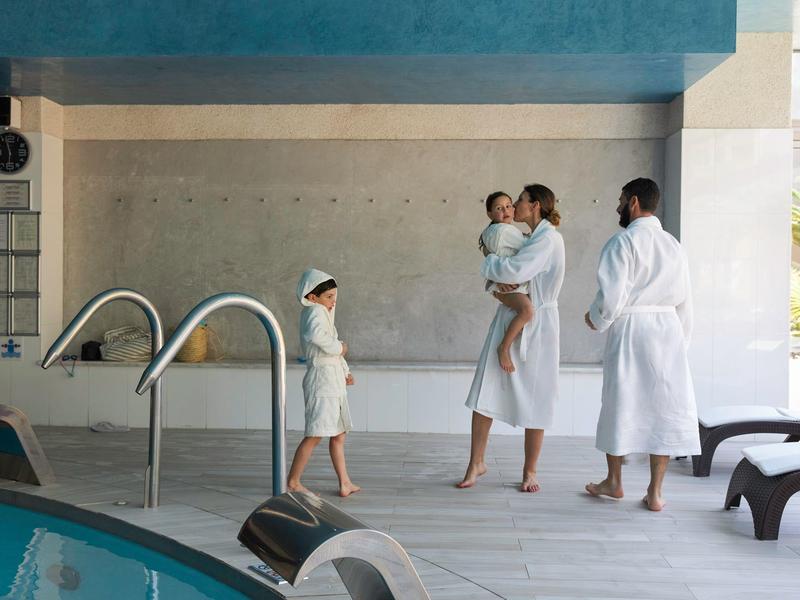 Trzy osoby w szlafrokach i dziecko stoją przy brzegu krytego basenu.