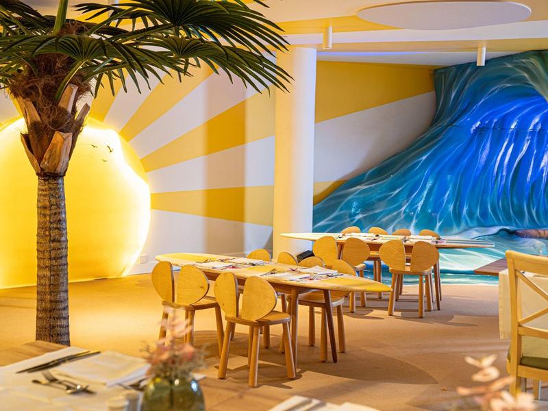 Restauracja z tropikalną dekoracją, palmami i muralem słońca i fali.