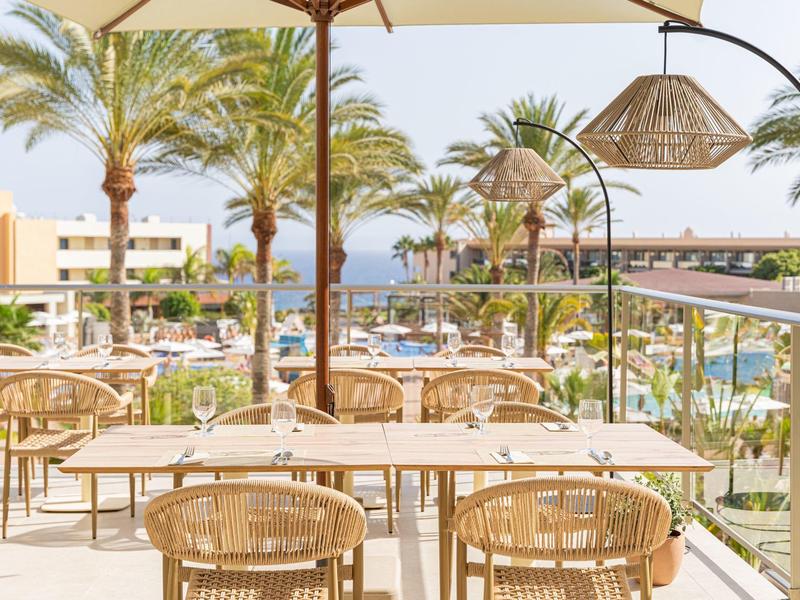 Widok na otwarty taras z stołami i krzesłami, palmami i basenem w tle.