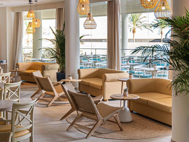 Jasna część wypoczynkowa z wygodnymi fotelami, sofami i dużymi oknami z widokiem na palmy.