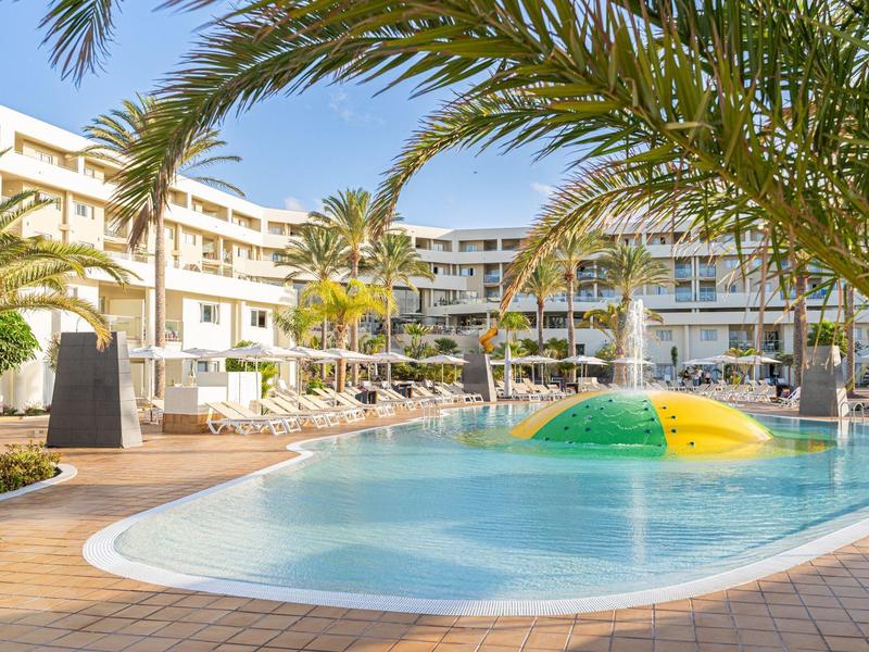 Hotel z basenem na zewnątrz, atrakcją wodną i palmami pod niebieskim niebem.