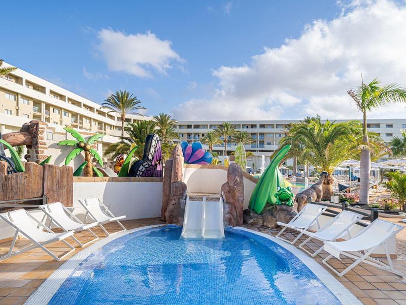 Basen hotelowy ze zjeżdżalnią wodną, leżakami i tropikalną dekoracją w słoneczną pogodę.