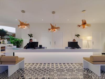 Moderne Rezeption mit weißem Tresen, zwei Computern, goldenen Hängelampen und gepolsterten Bänken.