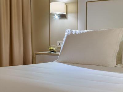 Helles, ordentlich gemachtes Bett mit weißer Bettwäsche neben beigem Vorhang und Wandlampe.