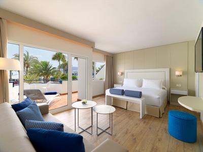 Helles Schlafzimmer mit großem Bett, blauer Sitzbank, Sofa, Holzboden und Terrasse mit Palmenblick.