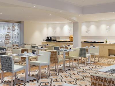 Bright hotel dining area with modern tables and chairs, carpeted floor, and counter in the background.