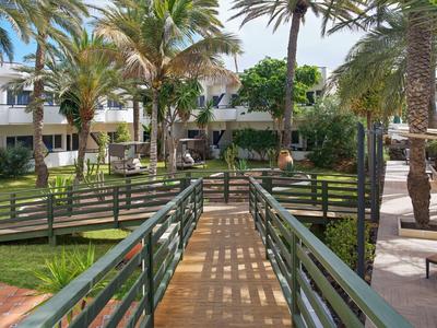 Green garden with palm trees and wooden bridge in front of a hotel building during daylight.