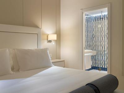 Simple hotel room with white bed, pillows, and adjoining bathroom with patterned shower wall.