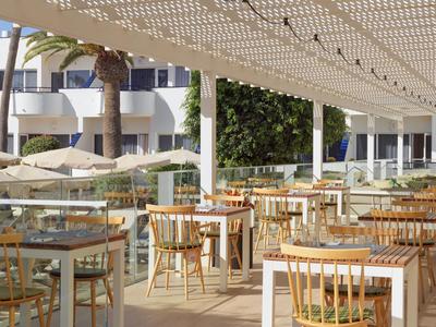 Hotel outdoor area with wooden chairs, white tables, and sunshade.