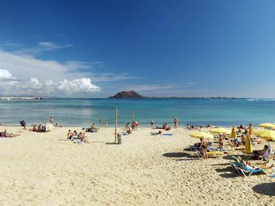 Strand mit hellem Sand, blauem Meer, Menschen und gelben Sonnenschirmen unter klarem Himmel.