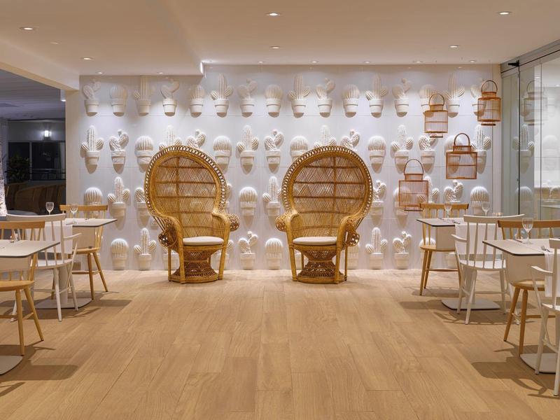 Área de restaurante moderna con piso de madera, sillas blancas y llamativos sillones de mimbre frente a una pared clara.