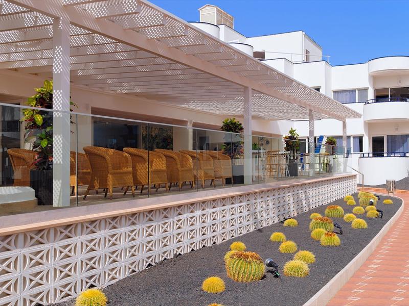 Terraza de hotel con sillas de mimbre bajo una pérgola y flores amarillas en primer plano