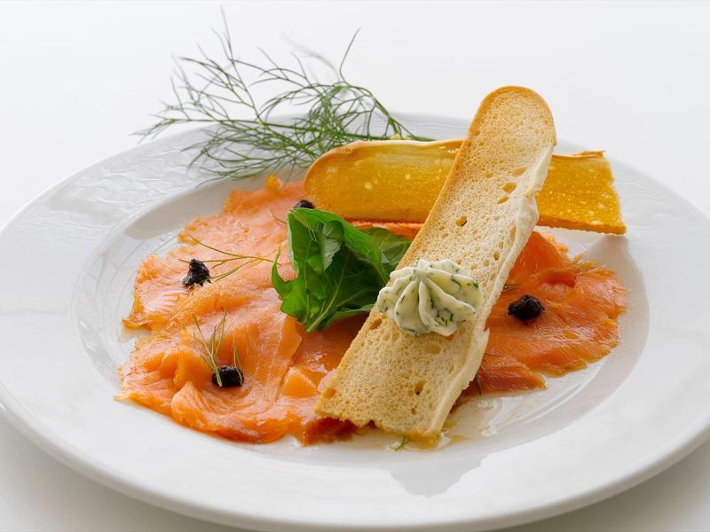 Plato con finas lonchas de salmón, tostada, hojas verdes y salsa amarilla en un plato blanco.