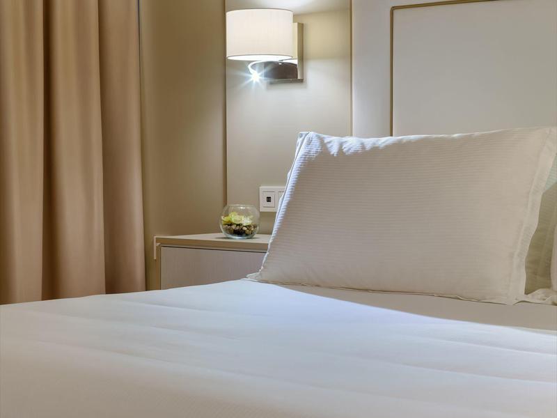 Habitación de hotel sencilla con cama blanca, almohada y lámpara de pared junto a cortinas beige.