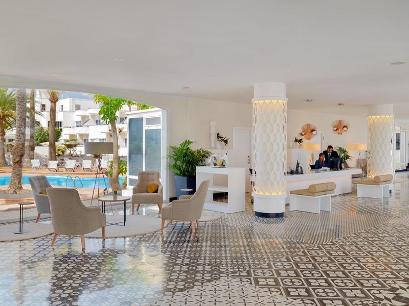 Vestíbulo de hotel luminoso con azulejos, áreas de descanso y vista a la piscina al fondo.