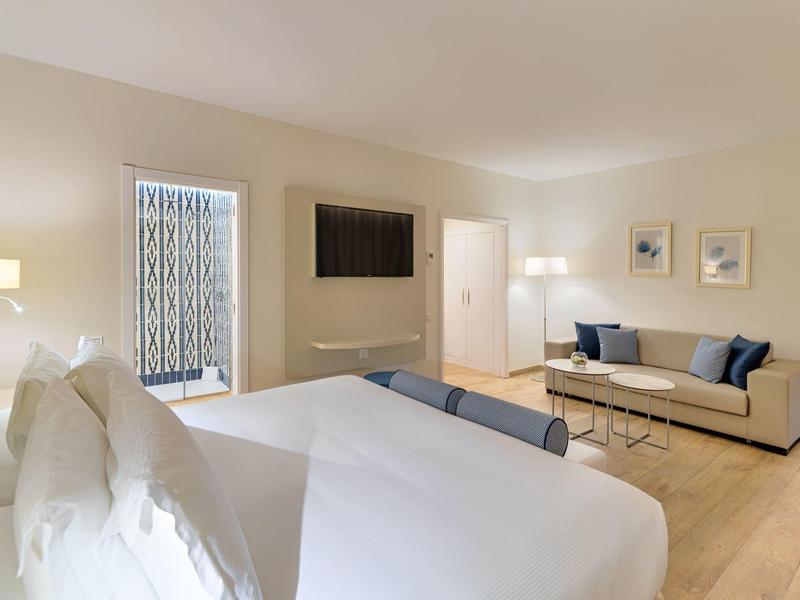 Habitación de hotel moderna con cama grande, sofás, televisor de pared y suelo de madera clara.