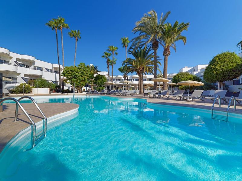 Piscina azul clara en un complejo hotelero con edificios blancos y palmeras bajo un cielo azul.