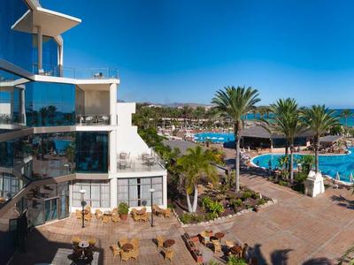 Complesso alberghiero moderno con piscina, palme e vista diretta sul mare sotto un cielo limpido.