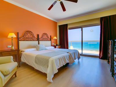 Camera d'hotel con due letti, parete arancione e vista mare tramite grandi finestre.