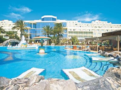 Großer Hotelpool mit Liegen, Palmen, Springbrunnen und weißem Hotelgebäude bei klarem Himmel.