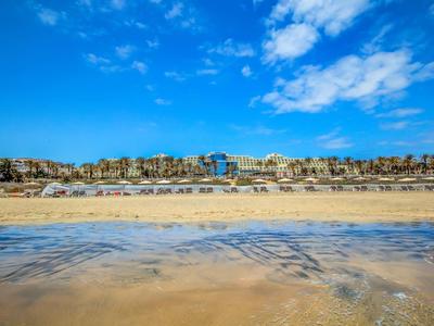 Ampia spiaggia sabbiosa con mare calmo davanti a una struttura alberghiera sotto un cielo azzurro.