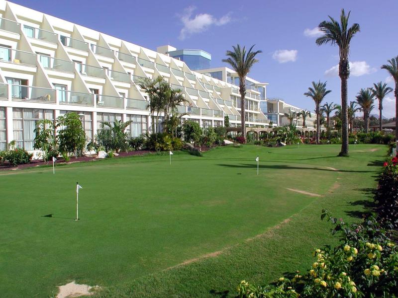 Ampio complesso alberghiero con area pratica golf curata e palme sotto il cielo azzurro.