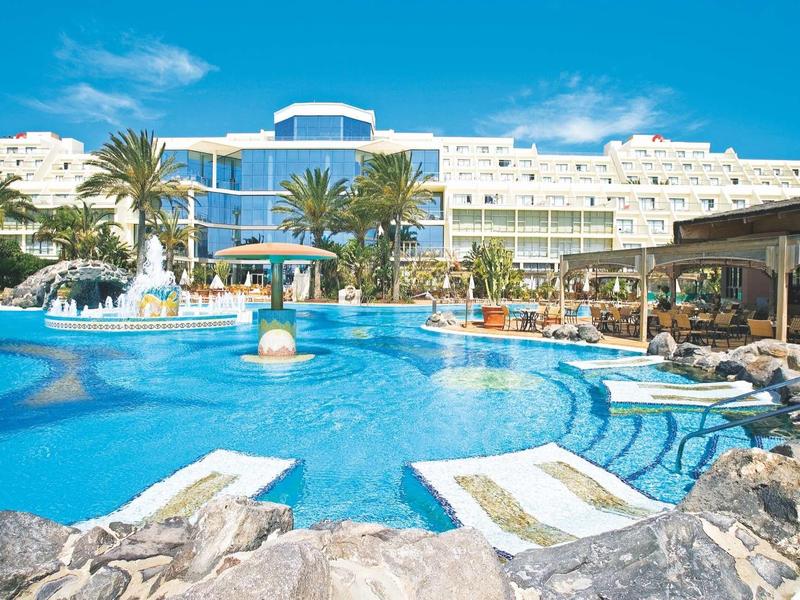 Hotel con grande piscina esterna, sdraio e palme sotto un cielo azzurro.