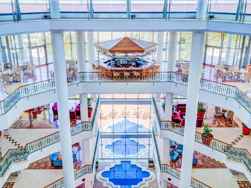 Atrio di hotel luminoso con galleria su più livelli, motivo di piastrelle blu e bar centrale