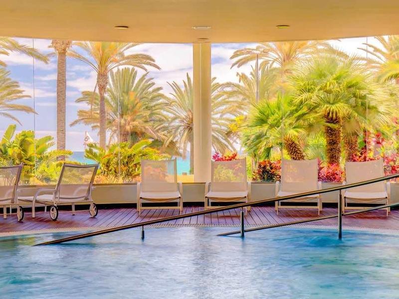 Piscina interna con grandi finestre che si affacciano su palme e mare, atmosfera tropicale dell'hotel