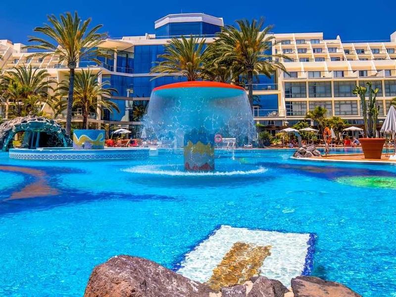 Grande piscina dell'hotel con fontana, lettini e palme sotto un cielo soleggiato.