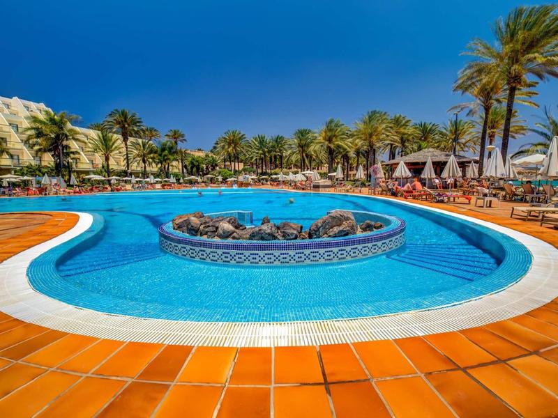 Grande piscina dell'hotel con ombrelloni, lettini e palme sotto un cielo blu sereno.